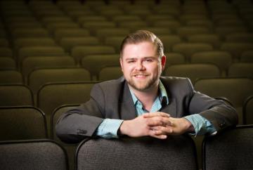 David wears a short beard, gray suitcoat with teal checked shirt, and is leaning over the back of seat in an empty theatre
