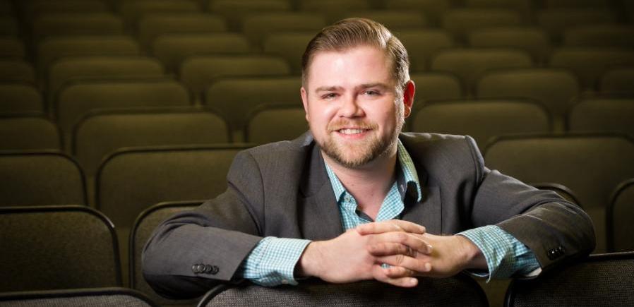 David wears a short beard, gray suitcoat with teal checked shirt, and is leaning over the back of seat in an empty theatre