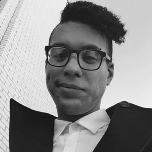 Black and white headshot of Patrick. He wears dark rimmed glasses, a white collared shirt with black overcoat and a smirk.