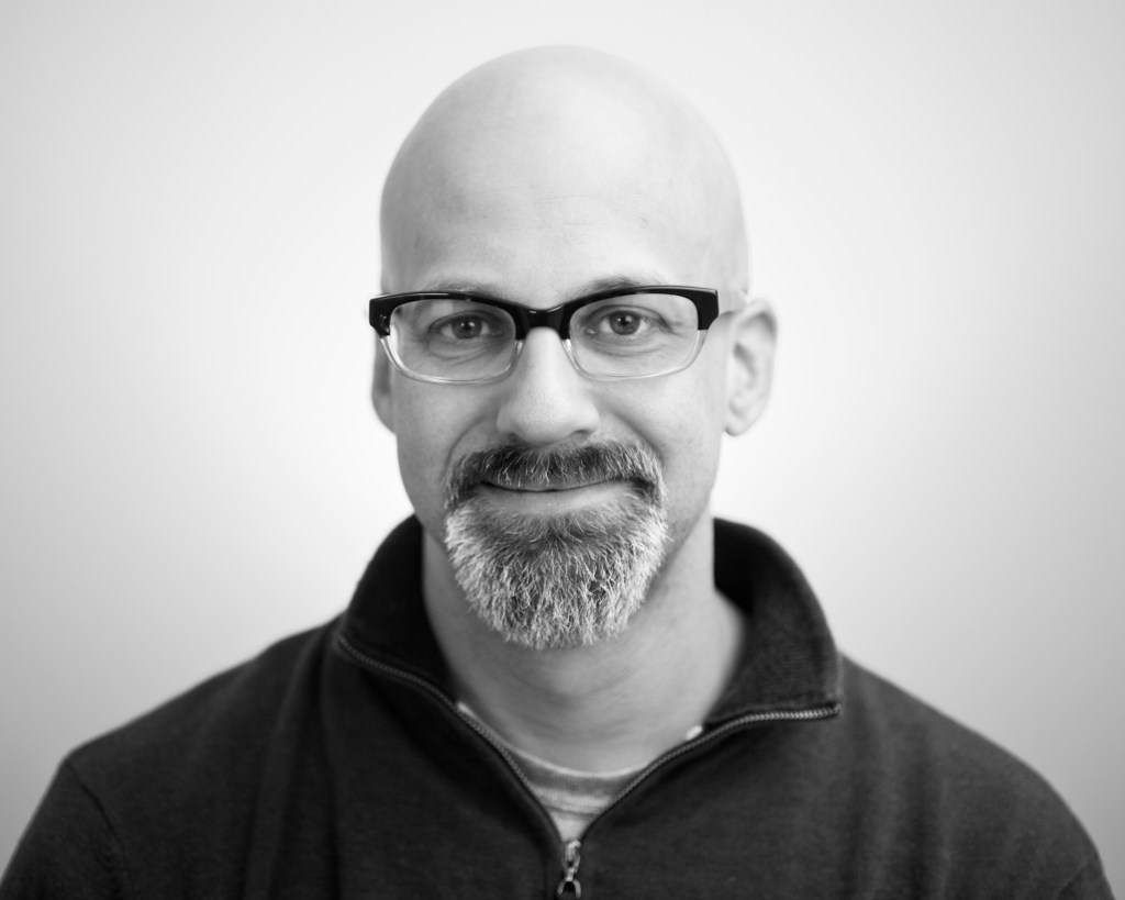 a lack and white headshot of Thaddeus. He wears dark rimmed glasses, a salt and pepper goatee, and a bald head with a dark collared collared zip up.